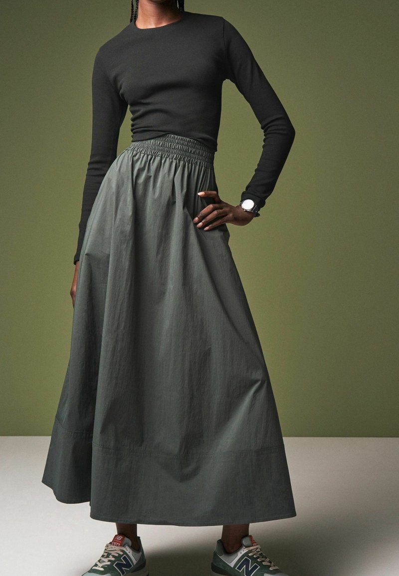 Femme portant un haut noir à manches longues, une jupe longue olive taille haute, une montre blanche au poignet et des baskets grises New Balance.