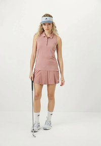 Egy nő rózsaszín ujjatlan pólót és szoknyát visel, kezében golfütővel, fehér Adidas napellenzőt, zoknit és sportcipőt hordozva egy sima fehér háttér előtt.