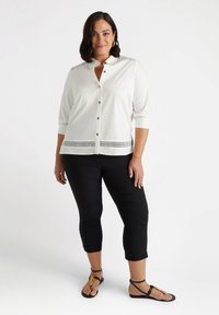 Blusa bianca con chiusura a bottoni frontale, maniche a tre quarti e inserti in rete decorativa all'orlo; abbinata a pantaloni neri a palazzo e sandali bassi.