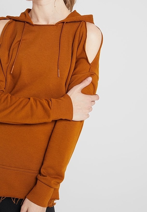Personne portant un sweat à capuche couleur rouille avec un détail ajouré à l'épaule, tenant son bras opposé devant un fond clair uni.