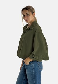 Olivgrön kort jacka med krage, knäppning framtill och vida ärmar. Har spänndetaljer på ärmslut och en slät textur.