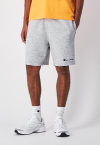Helle graue Baumwoll-Sportshorts mit einem elastischen Bund, seitlichen Taschen und einem kleinen rot-blauen Champion-Logo am linken Bein.