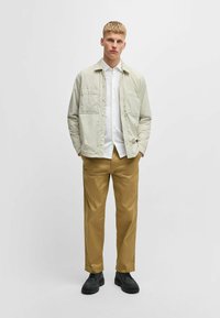 Veste matelassée beige clair avec deux poches poitrine, chemise blanche en dessous, pantalons khaki et chaussures noires épaisses. Design simple et structuré.