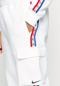 White Nike cargo pants feature red and blue stripe accents along the sides, with a large side pocket and a small logo on the front.