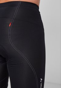 Leggings noirs avec des coutures blanches contrastantes, présentant une texture lisse, une taille haute et un petit détail de logo rouge sur le côté.