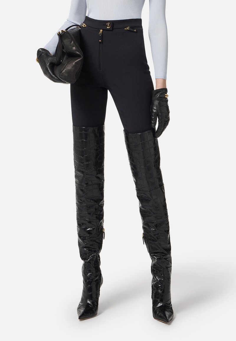 Leggings neri con vita alta realizzati in un tessuto liscio e dettagli con zip, abbinati a stivali alti fino alla coscia in nero lucido con effetto coccodrillo.