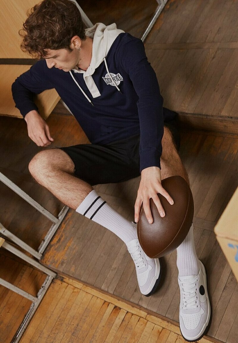 Granatowy hoodie z szarymi akcentami, czarne szorty, białe skarpetki w czarne paski i białe sneakersy, trzymając brązową piłkę nożną, siedząc na drewnianej podłodze.