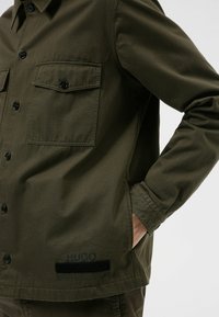 Chaqueta de botón en verde oliva, hecha de algodón, con dos bolsillos en el pecho, botones negros y un logo 'HUGO' en el dobladillo.