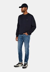 Felpa blu navy con tasca con zip sulla manica, abbinata a jeans azzurri e scarpe da ginnastica nere e bianche, tutto in uno stile casual e aderente.