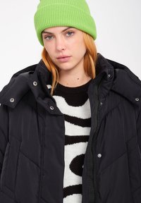Zwarte geïsoleerde jas met een rits aan de voorkant, drukknoopjes en een capuchon, gedragen over een zwart-wit gestreepte trui, gecombineerd met een felgroene beanie.