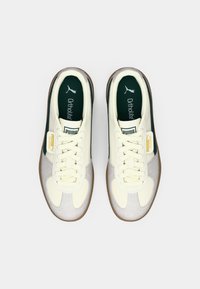 Par tenisica marke Puma u krem, sivoj i tamnozelenoj boji s bijelim vezicama i gumiranim potplatima, prikazanih odozgo na bijeloj pozadini.