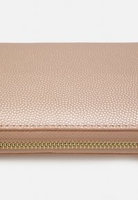 Portefeuille en rose gold avec une texture en cuir grainé, présentant une fermeture éclair dorée sur le bord. Forme rectangulaire compacte, doublure en tissu lisse.