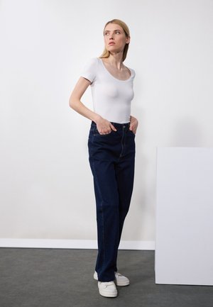 T-shirt alb mulat, blugi închiși la culoare cu talie înaltă și adidași albi. Modelul stă cu mâinile în buzunare pe un fundal neutru.