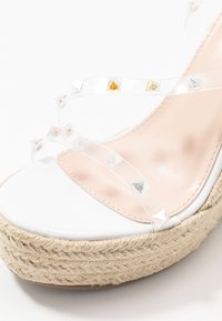 Sandale plate blanche avec une semelle en jute tissé. Dotée de brides transparentes ornées de clous triangulaires irisés et d'un semelle en nude.