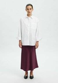Camicia bianca con bottone e tasca, abbinata a una lunga gonna di raso bordeaux e scarpe nere slip-on. Texture morbide e silhouette definite.