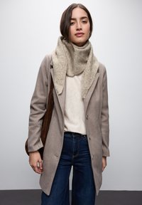 Manteau en laine beige clair au design ajusté, associé à une écharpe beige douce et un pull crème, complété par un jean bleu foncé.