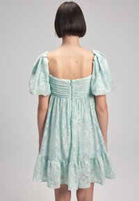 Vihreä minttuhame, jossa on puffihihat, kukkatekstuuri ja kerroksellinen röyhelöhelma. Takana on kerätty yksityiskohta, keskellä vetoketjulla.