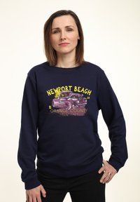 Felpa blu navy con grafica di una macchina vintage e testo "NEWPORT BEACH" in giallo. Tessuto morbido, maniche lunghe, polsini e orlo a costine.