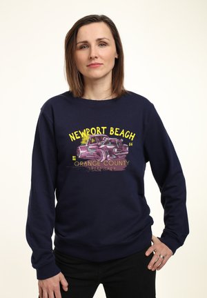 Sweatshirt bleu marine avec un graphique de voiture vintage et le texte "NEWPORT BEACH" en jaune. Tissu doux, manches longues, bords côtelés aux poignets et à l'ourlet.