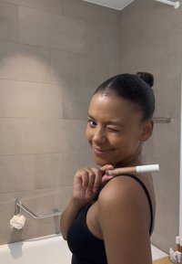 En concealer-stift med ett vitt lock hålls av en person som står i ett badrum. Väggarna har beige plattor och personen har på sig en svart linnen.