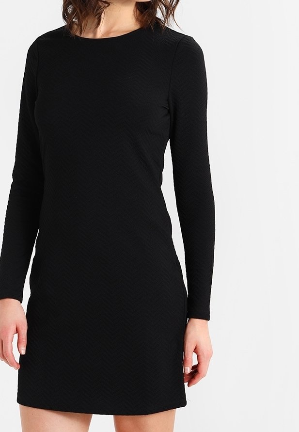 Femme portant une robe noire ajustée, à manches longues, longueur genou, debout devant un fond clair uni.