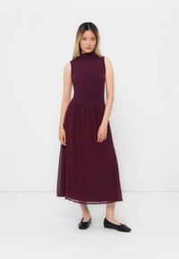 Young woman standing against white background wearing sleeveless high-neck dark purple midi dress and black ballet flats.