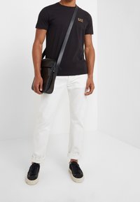 Black cotton t-shirt with gold logo on left chest, white denim pants, black textured messenger bag, and black sneakers with beige soles.