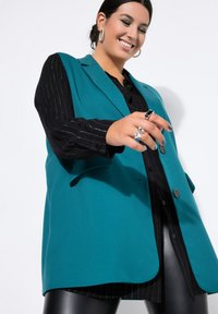 Blazer azul verdoso con un diseño estructurado, que presenta solapas con muesca y dos botones. Llevado sobre una camisa negra a rayas, combinado con leggings brillantes.