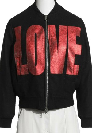 Veste noire zippée avec de grandes lettres rouges brillantes formant "LOVE" sur la poitrine, portée par-dessus un pantalon blanc.