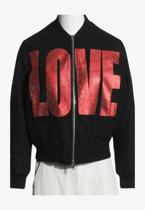 Veste noire zippée avec de grandes lettres rouges brillantes formant "LOVE" sur la poitrine, portée par-dessus un pantalon blanc.