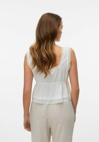 Top blanc sans manches avec bord en dentelle, légèrement froncé à la taille, présentant un design dos nu. Porté avec un pantalon de couleur claire.