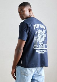 Tamsiai mėlyna medvilninė marškinėliai su grafiką ant nugaros, kurioje užrašyta "ŽAISK ŠALTAI" ir piešinys su personažu, laikydamu skateboard'ą.