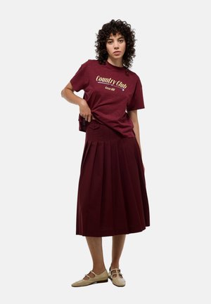 T-shirt bourgogne à manches courtes avec un graphique "Country Club" associé à une jupe plissée bourgogne. Le mannequin porte des chaussures beiges avec des découpes.
