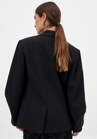 Čierny blazer so štruktúrovanými ramennými partami a jedným rozparkom na zadnej strane. Má dlhé rukávy a gombíkové akcenty na manžetách.