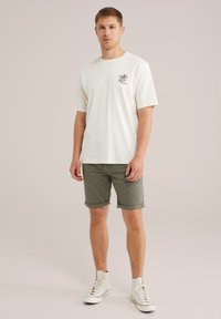 Nuori mies seisoo päällään valkoinen graafinen t-paita, oliivinvihreät käärityt shortsit ja valkoiset korkeat lenkkarit vaalealla taustalla.