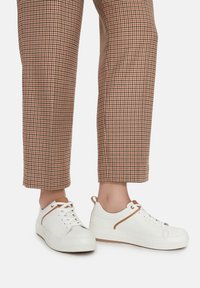 Białe sneakersy na sznurówki z teksturowaną powierzchnią i brązowym akcentem po bokach, zestawione z brązowymi spodniami w wzór houndstooth.