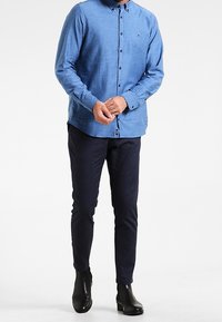 Man med en ljusblå långärmad skjorta, mörka slim-fit byxor och svarta ankelboots, stående mot en vit bakgrund.