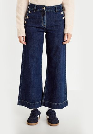 Kobieta nosząca ciemne, szerokie jeansy z szerokimi nogawkami i srebrnymi guzikami na biodrach, jasnoróżowy sweter oraz granatowe trampki z białymi paskami.