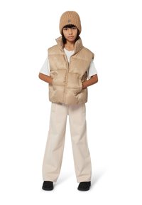 Beige puffer vest met een geometrisch patroon, gelaagd over een wit T-shirt. Beige wijde broek en een ribbelige beige beanie maken de outfit compleet.