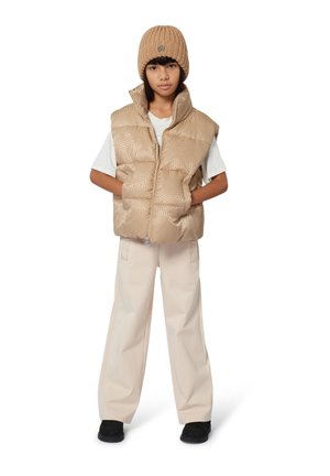 Beige puffer vest met een geometrisch patroon, gelaagd over een wit T-shirt. Beige wijde broek en een ribbelige beige beanie maken de outfit compleet.