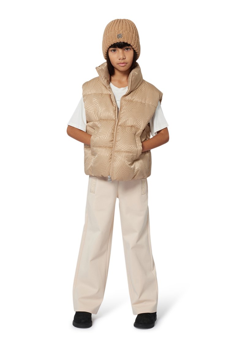Beige puffer vest met een geometrisch patroon, gelaagd over een wit T-shirt. Beige wijde broek en een ribbelige beige beanie maken de outfit compleet.