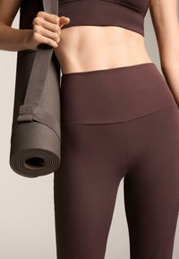 Hnědé legíny s vysokým pasem a odpovídající crop top vyrobené z hladké stretchy tkaniny; v pozadí je jóga podložka srolovaná a zajištěná popruhem.