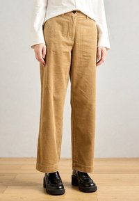 Persoon met beige corduroy broek, zwarte chunky hakloafers en een witte longsleeve, staand op een houten vloer tegen een effen achtergrond.