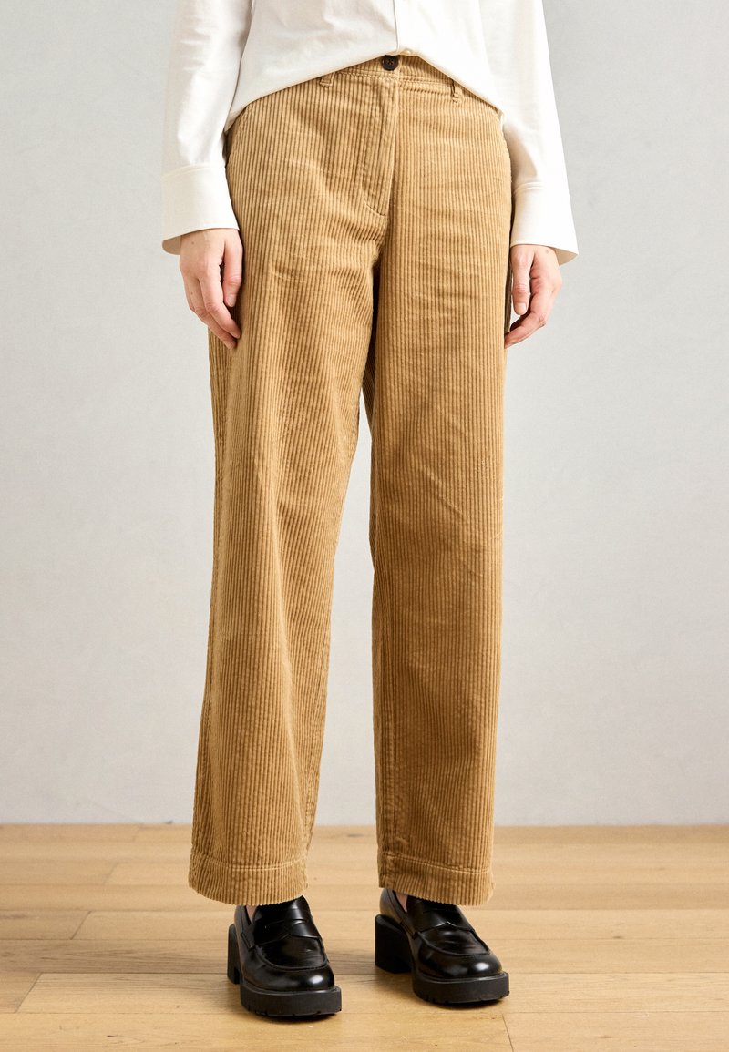 Persoon met beige corduroy broek, zwarte chunky hakloafers en een witte longsleeve, staand op een houten vloer tegen een effen achtergrond.
