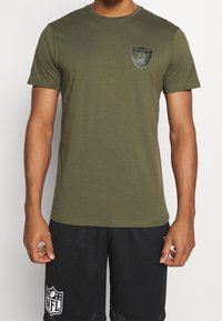 Olivgrön T-shirt med kort ärm av bomull, med en svart Raiders-logotyp på bröstet. Slät textur och rundad halsringning.