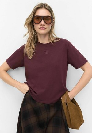 Femme aux cheveux blonds porte de grandes lunettes de soleil marron, un t-shirt bordeaux, une jupe à carreaux foncée, et tient un sac à main en daim marron sur fond blanc.