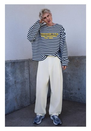 Persona giovane con capelli biondi corti che indossa una maglia a righe bianca e nera a maniche lunghe con testo giallo, pantaloni larghi color crema e scarpe da ginnastica grigie, in piedi.