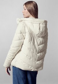 Giacca puffer in velluto a coste avorio con motivo a righe orizzontali, cappuccio e polsini elastici, abbinata a jeans di denim scuri.