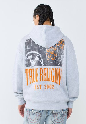 Person mit geflochtenem Haar trägt einen grauen Kapuzenpullover mit orange-schwarzer "True Religion Est. 2002" Grafik auf dem Rücken, kombiniert mit blauen Jeans.