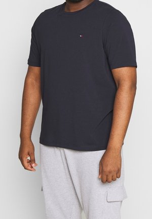 Mand iført sort kortærmet t-shirt med lille logo på brystet og lysegrå cargo bukser står foran en ensfarvet baggrund.